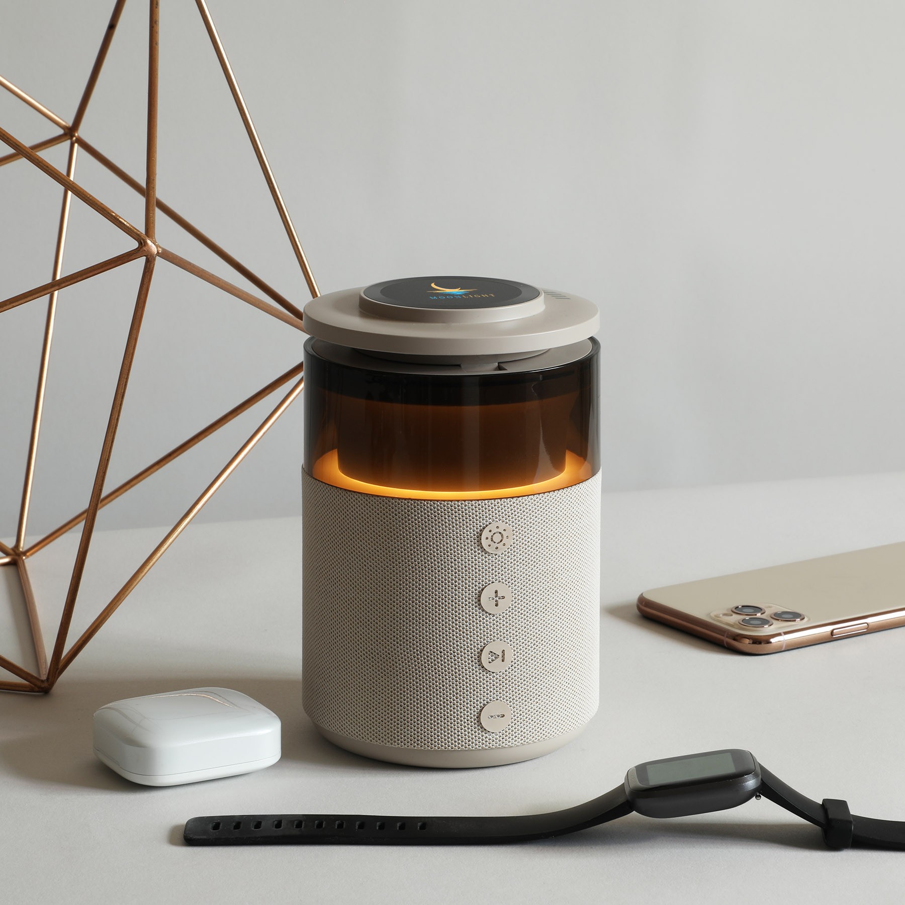 Cylindrical fabric speaker with a glowing amber light and a magnetic charger on top, beside earbuds, a phone, and a watch.