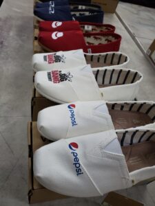 Row of white TOMS slip-on shoes, each decorated with a different custom logo